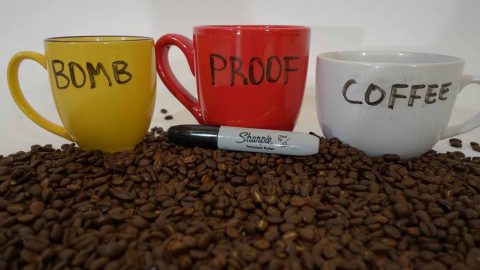 yellow cup, red cup, white cup spells out bomb proof coffee white background coffee beans foreground